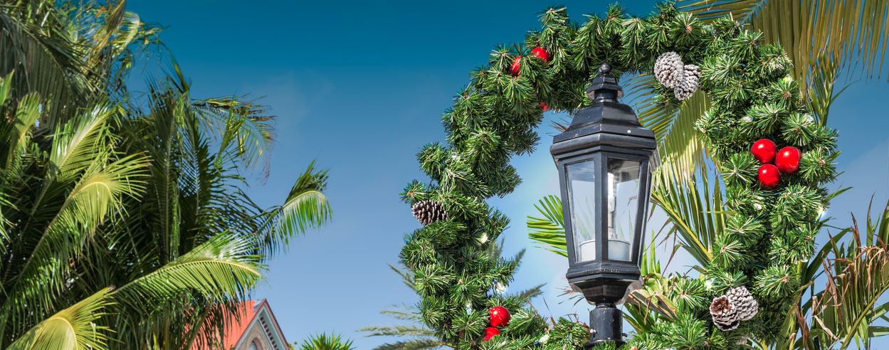 Holiday wreath with fir green, red baubles and pine cones, finished with a red bow. Christmas wreath around lantern lamp at a tropical vacation destination with palm trees in the Keys